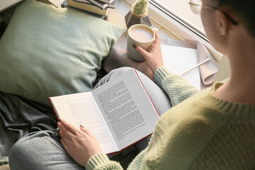 Woman with cup of coffee reading book near window, closeup