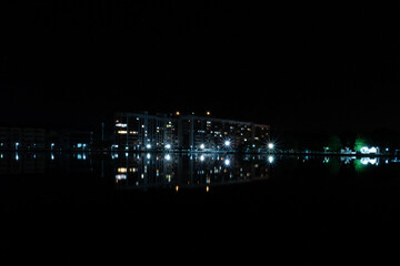 In the night water of the lake is reflected high-rise building with lighting. A nighttime cityscape. The street lights and the lights in the building windows are reflected in the water.