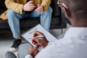 Close-up of psychotherapist making notes in patient card during conversation with patient at session - obrazy, fototapety, plakaty