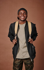 Portrait of African teenage boy with backpack smiling at camera standing against brown background