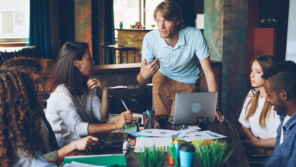 Enthusiastic CEO is talking to his team of professionals during meeting in loft style office while employees are listening to him and smiling. Teamwork and management concept.