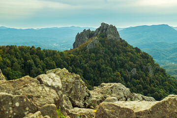 The rock rises above the green forest against the blue sky. A mountainous autumn landscape. Large rocks in the foreground. In the background are mountain peaks and passes.