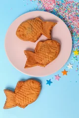 Poster Vertical top view of fish-shaped Taiyaki ice cream cones in a pink plate over the blue surface © Nina Ljusic/Wirestock Creators