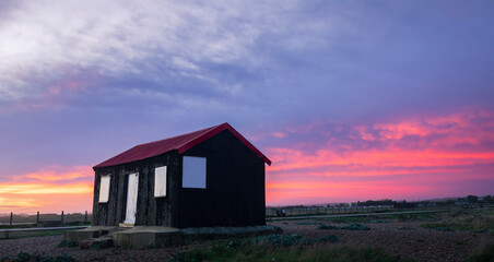 Obraz premium Stormy red skies at sunset over the red roofed hut Rye Harbour nature reserve on the East Sussex coast south east England UK