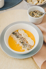 A white bowl with pumpkin soup garnished with pumpkin, sesame and sunflower seeds and crackers. In the background, in blur, pumpkin seeds in bowl. The table is covered with a linen tablecloth.