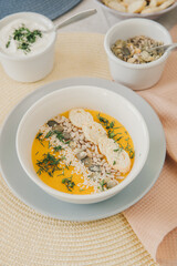 A white bowl with pumpkin cream soup garnished with pumpkin, sesame and sunflower seeds, crackers and dill. In the background, in blur, pumpkin seeds in bowl and sour cream in bowl.