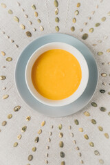Top view of white plate with pumpkin soup.  Peeled pumpkin seeds are arranged around the plate. The table is covered with a linen tablecloth. 