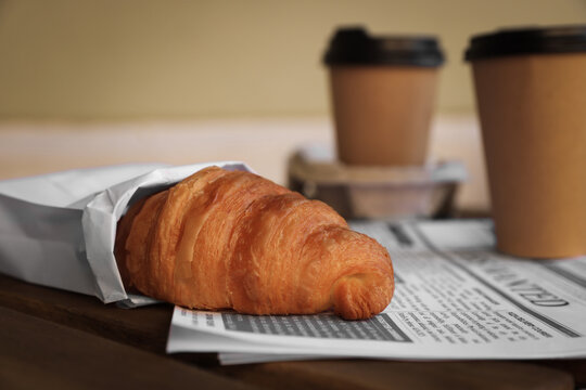 Tasty croissant, newspaper and paper cups of coffee on wooden table, closeup