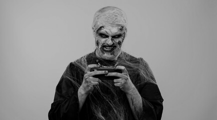 Sinister man with horrible scary Halloween zombie makeup enthusiastically playing racing drive video games on mobile phone. Dead guy with wounded bloody scars face isolated against gray background