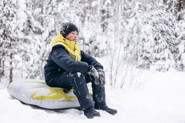Portrait of teenage boy sitting on tubing winter snowy forest. Child walking resting relaxing contemplating thinking daydreaming outdoors. Teen deep in thought