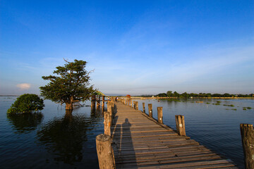 Old wooden bridge over calm lake water with gazebo and trees.