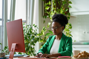 Successful African American woman working as editor business magazine typing article sits at computer desk. Girl with glasses performs remote work for Internet press located in home office with plants - obrazy, fototapety, plakaty