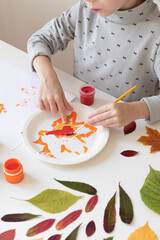 A little girl makes crafts on an autumn theme. She paints a birch leaf with red gouache to make a stamp. The girl got her fingers dirty. There is a white wall in the background.