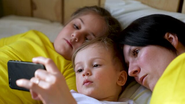 Happy family concept. Mom, daughter and son are play on bed. Child watching video on smartphone. Family leisure at home. Boy and girl with mom are watching movie on smartphone. Home education concept