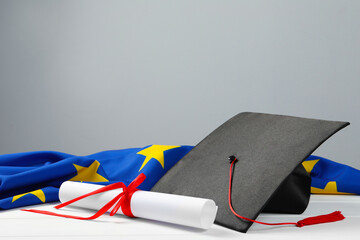 Black graduation cap, diploma and flag of European Union on white wooden table against grey wall