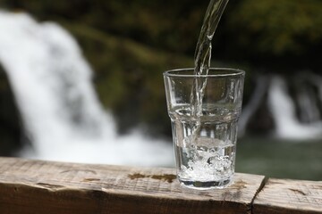 Fresh water pouring into glass on wooden surface near waterfall. Space for text
