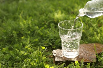 Pouring fresh water from bottle into glass on stone in green grass outdoors. Space for text