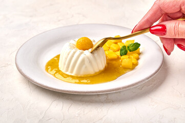 Woman's hand holds spoon over homemade panna cotta with natural vanilla pod, mango syrup and mint,...