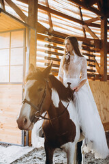 A charming boho bride rides a horse on a ranch at sunset in winter.