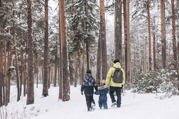 Rear view of father with backpack and little sons holding hand walking together in winter snowy forest. Wintertime activity outdoors. Concept of local travel and family weekend