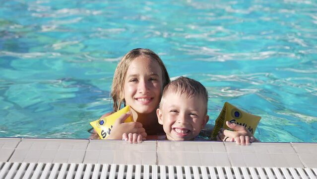 Close up of happy children swimming in pool. Brother and sister. Young beautiful girl and little boy laugh and smile broadly. Boy in inflatable armbands is holding onto side of pool. Look into camera.