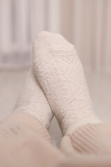 Women's feet in warm knitted woolen white socks at home under a plaid. Selective focus. Vertical photo.