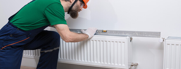 A male plumber installs a radiator in the heating system of an apartment. Guy in overalls and a gas...