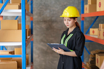 An Asian female factory worker in a reflective uniform with a hard hat inspects and records new arrivals in a warehouse.