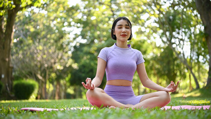 Beautiful Asian female in sportswear meditating on her yoga mat in green park.