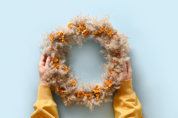 Woman holding in hands Thanksgiving wreath with orange flowers and dry natural materials over blue background. Top view.