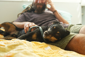 A bearded rastafarian hippie man petting his dog while talking on the phone, laying in his bed
