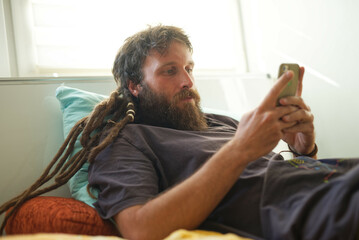 Young alternative fashion, reggae style man with dreadlocks, typing a message on his phone, laying...