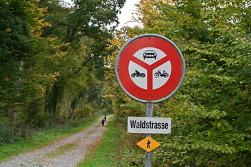 Road sign showing no entry for all motor vehicles. Below is signboard with text written in German languages Forest road and yellow sing with pictogram of hiker.