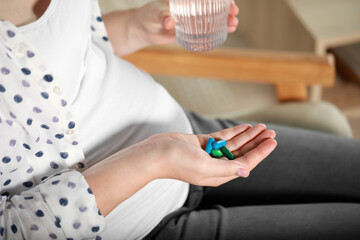 Pregnant woman holding pile of pills and glass with water indoors, closeup