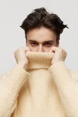 Vertical studio shot. a cute dark-haired young man stands in a beige turtleneck on a light gray background and smiles pleasantly, pointing his hands in different directions.