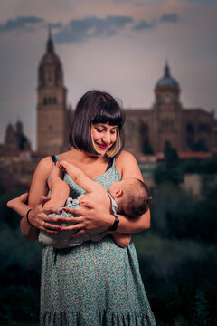 Joven madre con corte de pelo Bob y con un vestido largo estampado sujeta a su bebe en brazos el cual viste un overol de color azul verdoso. Atardecer de verano con la ciudad desenfocada de fondo 