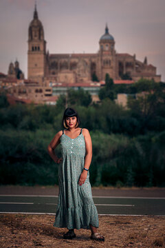Foto vertical de una mujer joven que modela con un vestido estampado azul verdozo, llevando un corte de pelo Bob. Atardecer en verano con la ciudad de fondo.