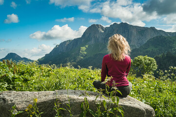 Naklejka premium A tourist girl in a purple sweater sits on a large rock against a mountain backdrop. Back view. High grass, blue sky with clouds. Travel and tourism. Mountain hike.