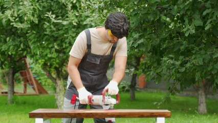 Young man builder carpenter grinds a wooden table with a grinding machine in the backyard. 