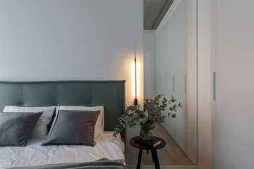 Modern minimalistic bedroom interior design in grey shades with light green bed, eucalyptus in glass vase, concrete ceiling. Lights on. Scandinavian style.  Aesthetic simple interior design concept.