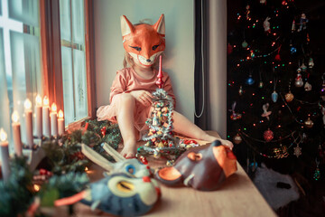 Cute little girl is dressing up a tiny Christmas tree and trying on New Year's animal masks. Image with selective focus and toning. 