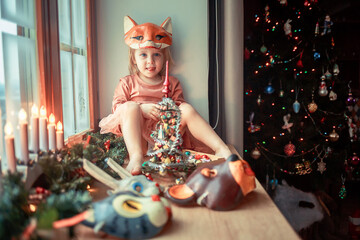 Cute little girl is dressing up a tiny Christmas tree and trying on New Year's animal masks. Image with selective focus and toning. 