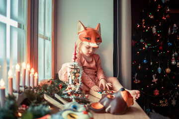 Cute little girl is dressing up a tiny Christmas tree and trying on New Year's animal masks. Image with selective focus and toning. 