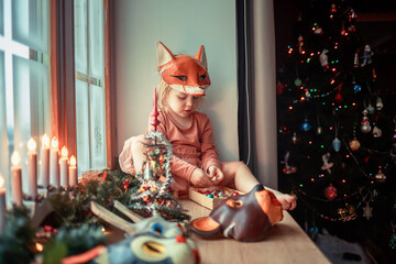 Cute little girl is dressing up a tiny Christmas tree and trying on New Year's animal masks. Image with selective focus and toning. 
