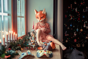 Cute little girl is dressing up a tiny Christmas tree and trying on New Year's animal masks. Image with selective focus and toning. 