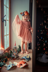 Cute little girl is dressing up a tiny Christmas tree and trying on New Year's animal masks. Image with selective focus and toning. 