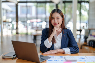 Portrait of smiling Asian businesswoman enjoying her hard work using her laptop at the office and looking at the camera.