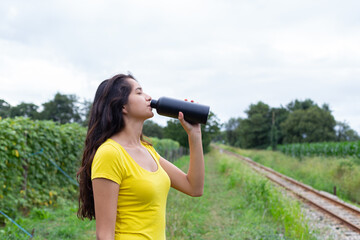 Young ethnic female athlete drinking water after training in nature 