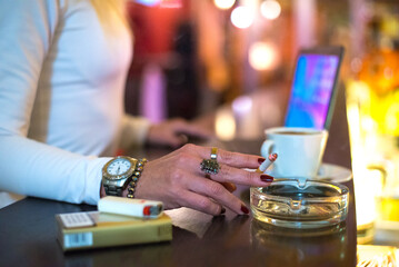 Middle aged blonde elegant woman sitting in the bar, drinking coffee and smoking cigarette while working on the laptop at night
