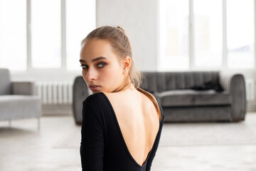 Portrait of young female in black outfit looking over the shoulder with sofa and window on background - 541452797